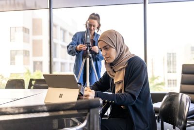 A woman sits at a table using a laptop, while another woman in the background adjusts a microphone on a tripod in a bright, modern room with large windows.